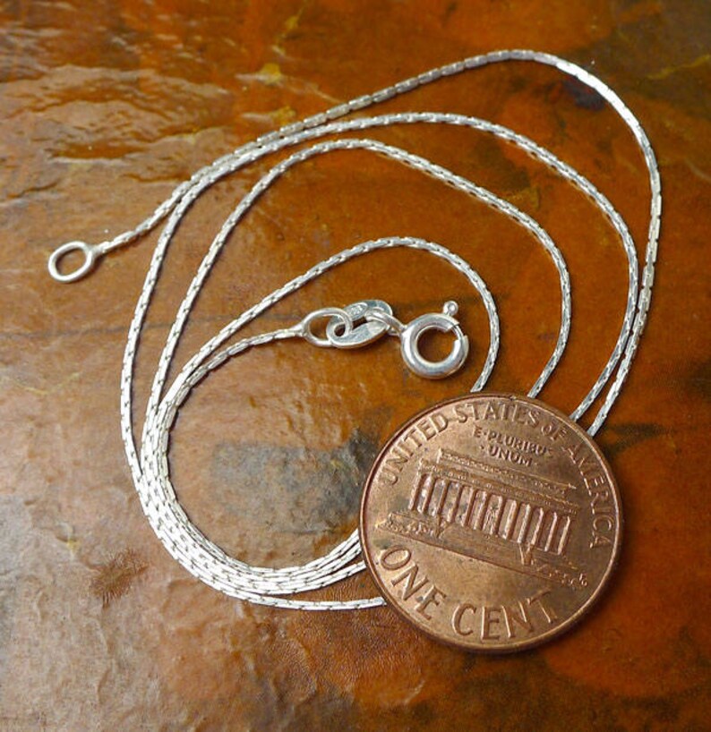 May include: A silver chain necklace with a lobster clasp. The chain is laid out on a brown surface next to a US penny.