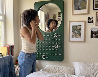 May include: A dark green wall-mounted calendar mirror featuring a circular reflective surface at the top and a grid of circular buttons below. The buttons are labeled with days of the week, numbers one through thirty-one, and months of the year.