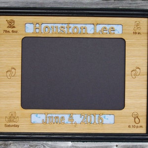 May include: A wooden picture frame with a black border and a cutout for a photo. The frame is decorated with a baby carriage, footprints, a teddy bear, and a rattle. The text "Houston Lee" is engraved on the top of the frame, and the text "June 4, 2016 6:10 p.m." is engraved on the bottom. The frame is 7 lbs. 6 oz. and 19 in. in size.