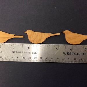 Può includere: Tre sagome di uccelli in legno, ciascuna lunga circa 5 cm, sono disposte in fila su una superficie nera. Un righello è posizionato sotto gli uccelli per mostrare le loro dimensioni.
