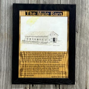 May include: A framed wall art piece featuring a black wooden frame, a central sketch of a barn labeled The Mule Barn, and a detailed historical narrative engraved on a light wood background describing the history of the Mule Barn in Sheridan.
