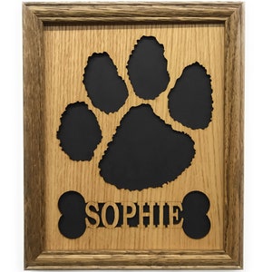May include: A rectangular wooden frame containing a light brown wood cutout of a large animal paw print and a bone shape with the name SOPHIE centered in the middle. The cutout is layered over a solid black background, creating a high contrast.