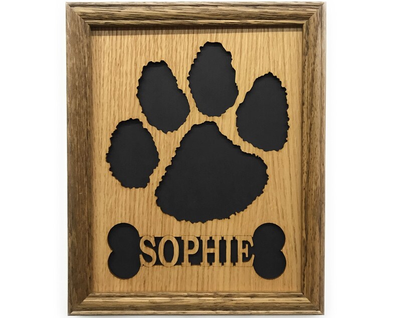 May include: A rectangular wooden frame containing a light brown wood cutout of a large animal paw print and a bone shape with the name SOPHIE centered in the middle. The cutout is layered over a solid black background, creating a high contrast.