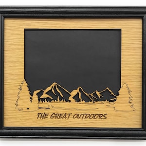 May include: A framed wooden art piece with a black frame and a light brown inner frame. The artwork features a mountain and tree landscape cut-out design with the words "THE GREAT OUTDOORS" at the bottom.