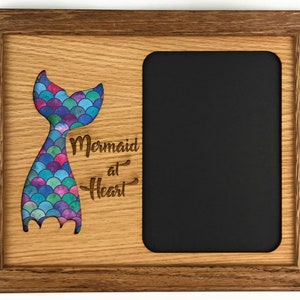 Puede incluir: Un marco de fotos de madera con un recorte de una cola de sirena con escamas de colores. El texto "Mermaid at Heart" está grabado debajo de la cola. El marco tiene un paspartú negro y un acabado de madera marrón.