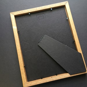 May include: A rectangular picture frame with a light brown wooden border and a black backing. The frame includes a black easel stand attached to the back, along with several small metal tabs used to secure the backing in place within the frame.