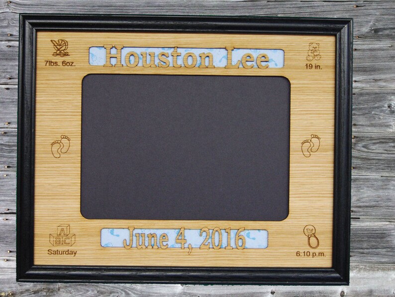 May include: A wooden picture frame with a black border and a cutout for a photo. The frame is decorated with a baby carriage, footprints, a teddy bear, and a rattle. The text "Houston Lee" is engraved on the top of the frame, and the text "June 4, 2016 6:10 p.m." is engraved on the bottom. The frame is 7 lbs. 6 oz. and 19 in. in size.