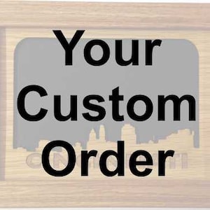 May include: A light-colored wooden picture frame with the words "Your Custom Order" in bold black letters. The background is a gray rectangle with a silhouette of a cityscape. The frame has a simple, classic design.