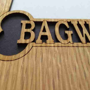 May include: A light brown wooden sign featuring a laser-cut design with the text BAGWE visible. The letters are carved out of the wood, revealing a dark background behind the cutout, showcasing the natural grain and texture of the wood surface.
