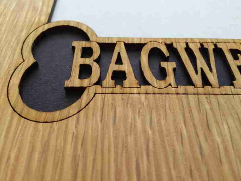May include: A light brown wooden sign featuring a laser-cut design with the text BAGWE visible. The letters are carved out of the wood, revealing a dark background behind the cutout, showcasing the natural grain and texture of the wood surface.