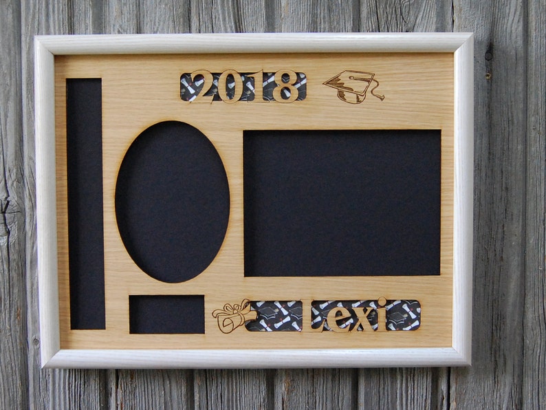 Peut inclure: Un cadre photo de remise de dipl&ocirc;me en bois avec une bordure blanche. Le cadre comporte des espaces pour des photos de diff&eacute;rentes formes, dont un ovale et un rectangle. L'ann&eacute;e "2018", une toque de dipl&ocirc;m&eacute; et le nom "Lexi" sont d&eacute;coup&eacute;s dans le bois.