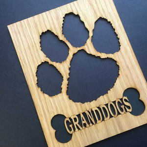 May include: A rectangular light brown wooden plaque featuring a laser-cut paw print design at the top and a bone-shaped cutout at the bottom. The word GRANDDOGS is engraved in capital letters across the center of the bone-shaped cutout area.