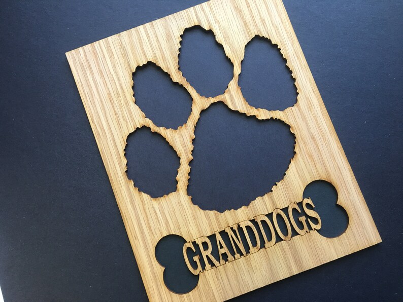 May include: A rectangular light brown wooden plaque featuring a laser-cut paw print design at the top and a bone-shaped cutout at the bottom. The word GRANDDOGS is engraved in capital letters across the center of the bone-shaped cutout area.