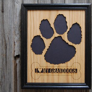 May include: A rectangular wall frame with a black border containing a light brown wood panel. The panel features a large cutout in the shape of a paw print revealing a black background and a bone-shaped cutout with the text I LOVE MY GRANDDOGS.
