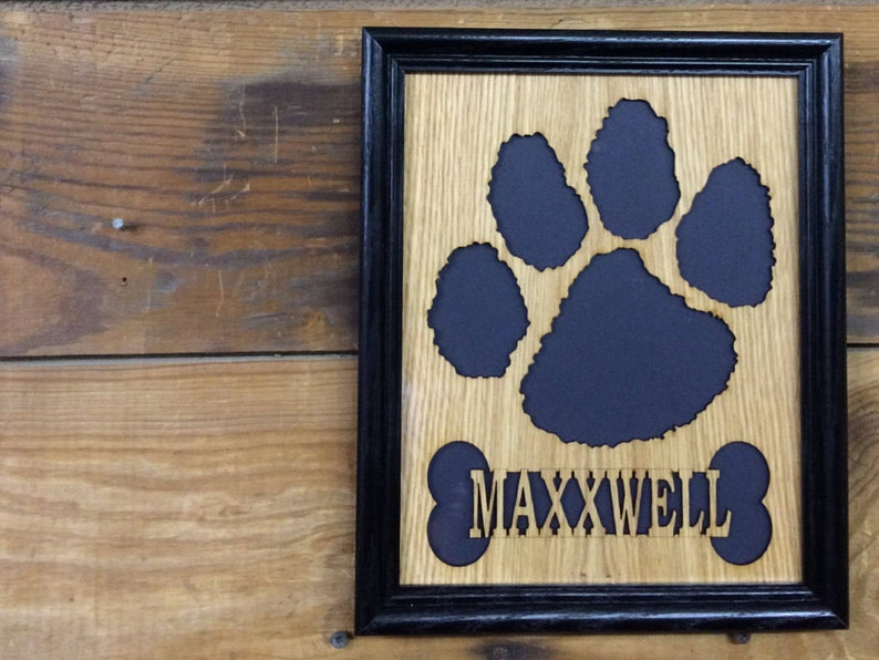 May include: A rectangular wall plaque in a black frame featuring a light brown wood grain background. A dark cutout of a paw print sits above a dark bone-shaped cutout with the name MAXXWELL in light wood lettering centered on the bone.