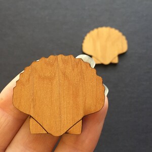 Può includere: Due forme di conchiglie in legno, una in una mano e l'altra su una superficie nera. Le conchiglie sono di colore marrone chiaro e hanno una texture a venature di legno.