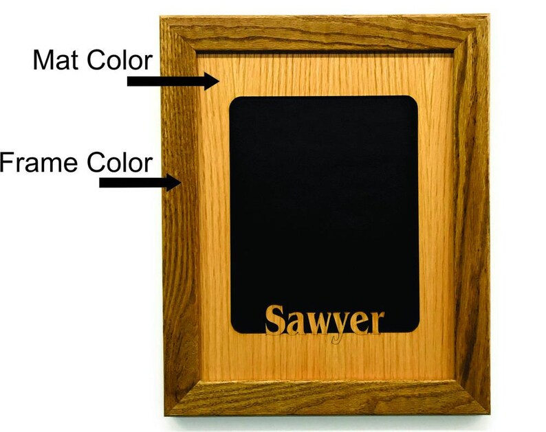May include: A rectangular picture frame with a dark brown wood grain finish and a light tan wood grain mat. The center features a solid black rectangular insert with the name Sawyer printed in a gold-colored serif font at the bottom center.