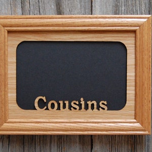 Puede incluir: Un marco de fotos rectangular de madera con un interior negro y la palabra "Cousins" recortada en letras de madera. El marco es de madera marrón claro y está sobre un fondo de madera envejecida.