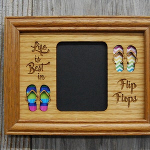 Puede incluir: Marco de fotos de madera marrón claro con la inscripción grabada "Life is Best in Flip Flops". El marco presenta diseños de chanclas coloridas y una abertura central para una foto.