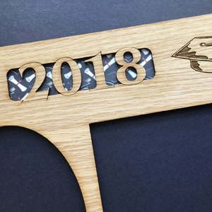 Peut inclure: Cadre de dipl&ocirc;me en bois avec l'ann&eacute;e "2018" d&eacute;coup&eacute;e, r&eacute;v&eacute;lant un tissu &agrave; motifs. Un chapeau de graduation est grav&eacute; &agrave; droite. Le cadre a une ouverture circulaire et rectangulaire pour les photos. Le fond est sombre.