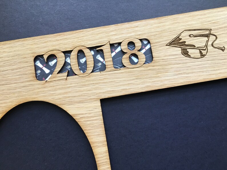 Peut inclure: Cadre de dipl&ocirc;me en bois avec l'ann&eacute;e "2018" d&eacute;coup&eacute;e, r&eacute;v&eacute;lant un tissu &agrave; motifs. Un chapeau de graduation est grav&eacute; &agrave; droite. Le cadre a une ouverture circulaire et rectangulaire pour les photos. Le fond est sombre.
