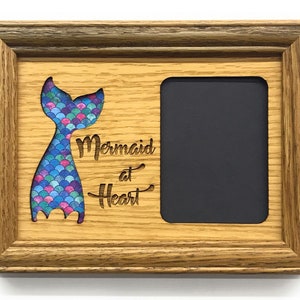 Puede incluir: Un marco de fotos de madera con un recorte de una cola de sirena con escamas azules, rosas y verdes. El texto "Mermaid at Heart" está grabado debajo de la cola. El marco tiene un paspartú negro y un cristal transparente.