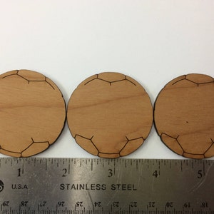 Può includere: Tre sottobicchieri in legno a forma di pallone da calcio, ognuno con un diametro di circa 5 cm. I sottobicchieri sono in legno marrone chiaro e hanno un contorno marrone scuro a forma di pallone da calcio.