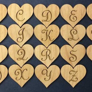 May include: A collection of twenty-six light brown wooden heart-shaped cutouts, each featuring a single engraved letter of the alphabet in a cursive script font. The hearts are arranged in four rows, displaying the letters A through Z in order.