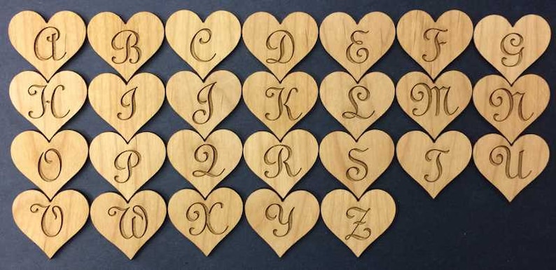 May include: A collection of twenty-six light brown wooden heart-shaped cutouts, each featuring a single engraved letter of the alphabet in a cursive script font. The hearts are arranged in four rows, displaying the letters A through Z in order.