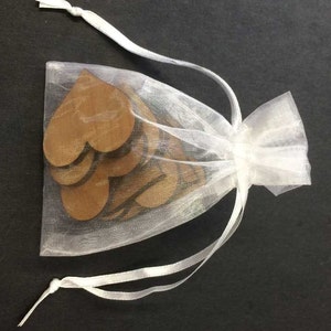 May include: A small, semi-transparent white organza drawstring bag containing several flat, brown, heart-shaped wooden cutouts. The bag is cinched at the top with two white ribbon ties, allowing the wooden heart shapes to be partially visible inside.