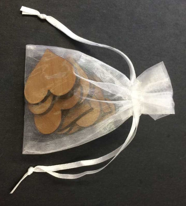 May include: A small, semi-transparent white organza drawstring bag containing several flat, brown, heart-shaped wooden cutouts. The bag is cinched at the top with two white ribbon ties, allowing the wooden heart shapes to be partially visible inside.