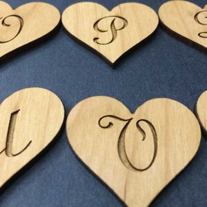 May include: A collection of light brown wooden heart-shaped cutouts, each featuring a single engraved letter in a cursive script. The visible letters include O, P, Q, U, and V, with each piece displaying a smooth, natural wood grain texture.