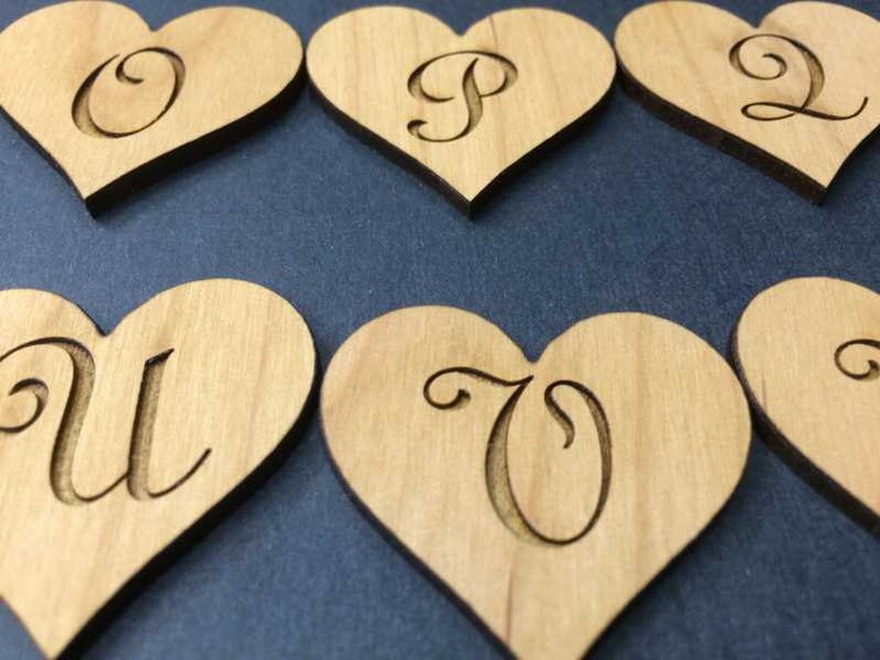 May include: A collection of light brown wooden heart-shaped cutouts, each featuring a single engraved letter in a cursive script. The visible letters include O, P, Q, U, and V, with each piece displaying a smooth, natural wood grain texture.