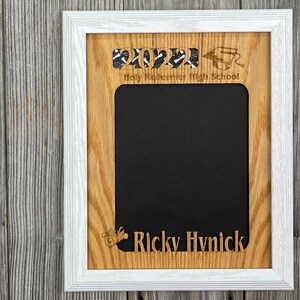 May include: A white wood picture frame with a brown wood mount. The frame has a cutout of the year 2022, a graduation cap, and the text "Holy Redeemer High School". The bottom of the frame has a cutout of a graduation cap and the name "Ricky Hvnick".