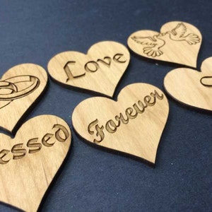 May include: Six light brown wooden heart-shaped cutouts featuring laser-engraved designs. The engravings include two interlocking rings, the words Love, Blessed, Forever, a pair of doves, and a stylized letter S, all displayed on a dark surface.
