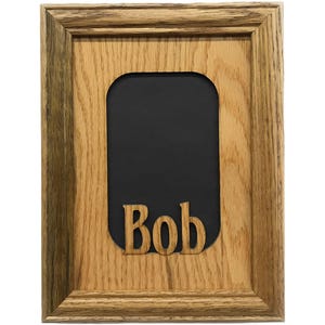 Puede incluir: Marco de fotos de madera con forma rectangular y acabado de roble marrón claro. El marco tiene un interior negro con el nombre "Bob" recortado en letras de madera.