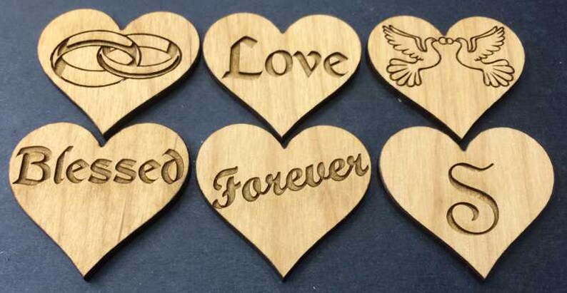May include: Six light brown wooden heart-shaped cutouts arranged in two rows. The top row features engraved images of two interlocking rings, the word Love, and two doves. The bottom row features the words Blessed, Forever, and the letter S.