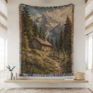 May include: A woven throw blanket featuring a detailed landscape scene of a rustic log cabin nestled in a dense pine forest at the base of snow-capped mountains, with a wildflower meadow in the foreground and a fringed edge on all four sides.