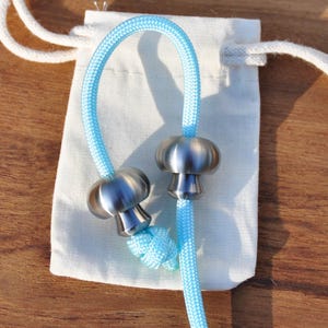 May include: A light blue nylon cord featuring two metallic, mushroom-shaped beads with a smooth, polished finish. The cord is looped and rests atop a small, off-white fabric drawstring pouch with white cotton strings tied at the top.