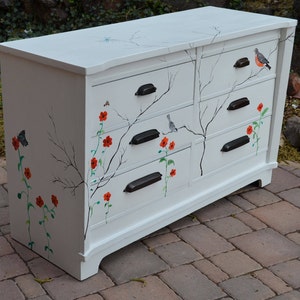 May include: White dresser with six drawers painted with a whimsical nature scene. The drawers feature black handles and are decorated with branches, birds, and red flowers.