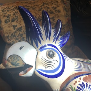 May include: A ceramic parrot figurine with blue and white painted details. The parrot has a white beak and a black eye. The figurine is painted in a traditional Mexican style.
