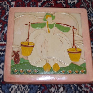 May include: A ceramic tile with a painted image of a woman in a white dress carrying two buckets. The woman is standing on a green hill with a windmill in the background. The tile has a pink border.