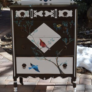 May include: A vintage dresser with a dark brown finish and white accents. The dresser has four drawers and is decorated with painted birds and branches. The dresser is on a brick patio.