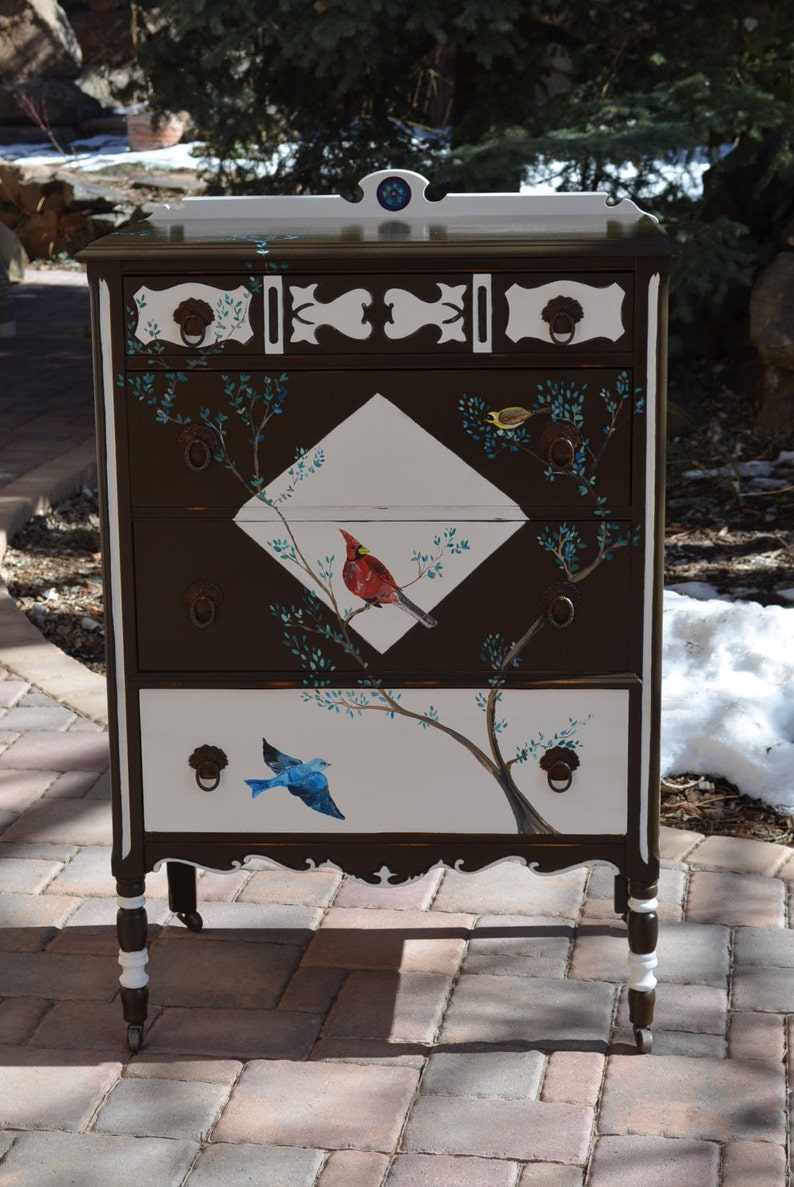 May include: A vintage dresser with a dark brown finish and white accents. The dresser has four drawers and is decorated with painted birds and branches. The dresser is on a brick patio.