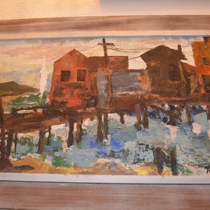 May include: Oil painting of waterfront buildings on stilts, with a muted color palette of reds, browns, and blues. The artwork is framed in a light-colored wooden frame, showcasing a vintage aesthetic.