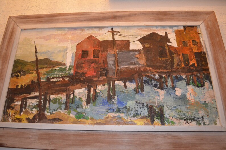 May include: Oil painting of waterfront buildings on stilts, with a muted color palette of reds, browns, and blues. The artwork is framed in a light-colored wooden frame, showcasing a vintage aesthetic.