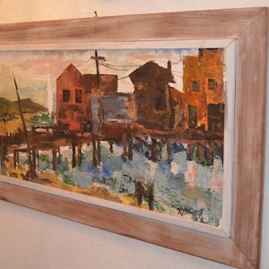 May include: Oil painting of a waterfront scene with buildings in shades of red, brown, and orange, reflected in the water. The artwork is framed in a light-colored wooden frame, creating a rustic aesthetic. The painting depicts a coastal landscape.