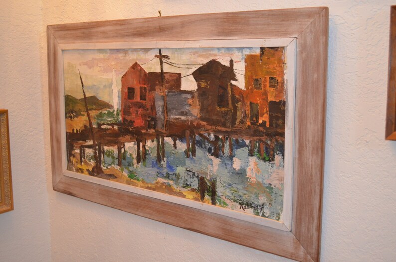 May include: Oil painting of a waterfront scene with buildings in shades of red, brown, and orange, reflected in the water. The artwork is framed in a light-colored wooden frame, creating a rustic aesthetic. The painting depicts a coastal landscape.