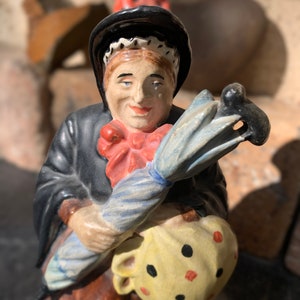 May include: A ceramic figurine of a woman wearing a black hat and dress, holding a blue and white striped umbrella and a yellow bag with red and black polka dots.
