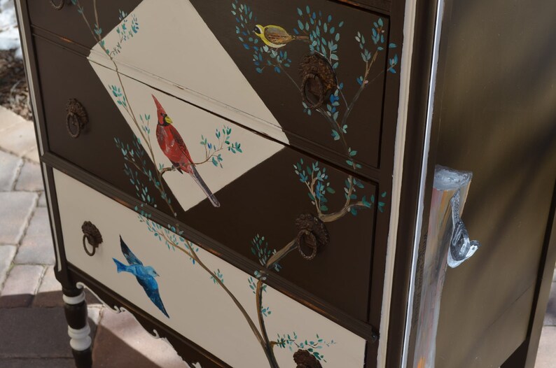 May include: A brown dresser with three drawers, each painted with a different bird and floral design. The top drawer features a yellow bird, the middle drawer has a red cardinal, and the bottom drawer has a blue bird. The dresser has black metal drawer pulls.